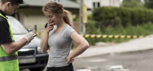 A uniformed officer takes notes while a woman stands with her hand on her chin, appearing concerned, at a roadside scene.