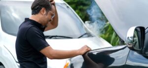 A man in a black shirt looks stressed as he checks the engine of a car, with smoke rising from under the hood.