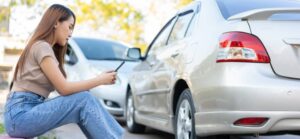 A person sits casually on the ground, looking at their phone, with parked cars in the background and trees softly blurred.