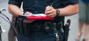 A police officer in a uniform holding a red clipboard, writing notes, with a bicycle nearby and blurred pedestrians in the background.