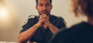 A police officer sits at a table with hands clasped, engaged in a serious conversation, with a blurred figure across from him.