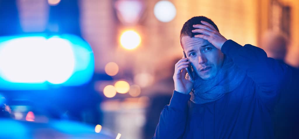 A person in a dark jacket holds their head with one hand, standing near a police car with flashing blue lights in a nighttime scene.
