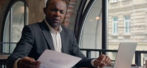 A man in a suit sits at a cafe table, holding a document while a laptop is open in front of him, with large windows in the backdrop.