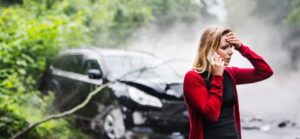 A woman stands in distress by a black car that has crashed into a wooded area, enveloped in fog and foliage.