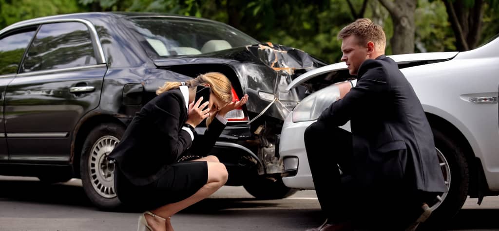 A woman in a business suit is on the phone, distressed, near two damaged cars after an accident, while a suited man kneels beside her.
