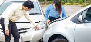 Two individuals inspect their cars after a minor accident, with visible damage on the front of both vehicles in a parking area.
