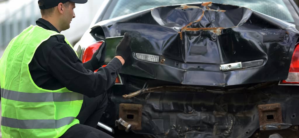 A person in a reflective vest inspects the damaged rear of a black car, showing signs of a serious collision.